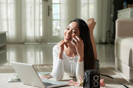 Smiling attractive Asian girl lying on carpet with laptop and vintage camera and talking on phone while working with photosの写真素材