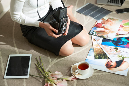 Close-up of unrecognizable female photographer in black skirt sitting on knees with vintage camera among bright colorful photos on carpetの写真素材