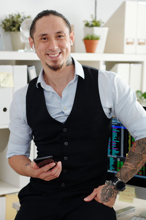 Portrait of handsome smiling trader standing at his table with smartphone in hands and looking at...