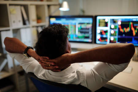 Relaxed trader leaning back in his chart and looking at ascending chart on computer monitors in front of himの写真素材