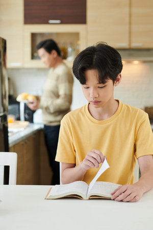 Serious teenage boy reading book when his father cooking breakfast in background