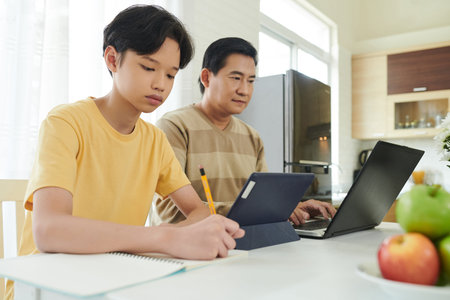 Concentrated teenage boy doing homework at kitchen table when sitting next to his father working on...