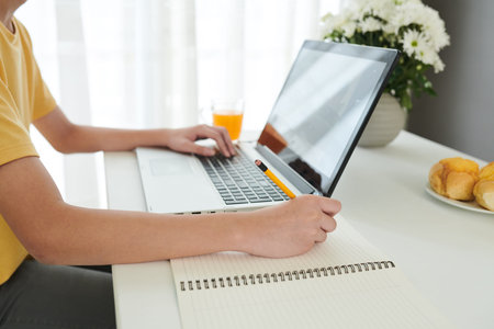 Hands of schoolboy reading book or article on laptop and writing in textbook at desk in his room