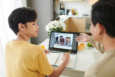 Father controlling teenage son watching online webinar on laptop when sitting at kitchen table at...