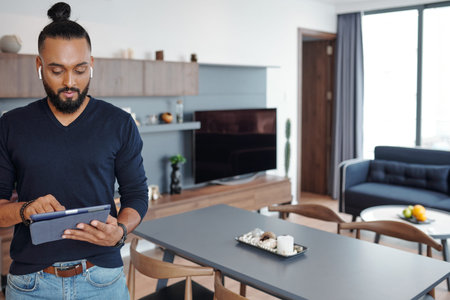 Man with wireless earbuds standing in his apartment and choosing music on tablet computerの写真素材