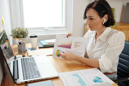 Businesswoman pointing at diagram in her hands when explaining marketing trends to her team during online meetingの写真素材