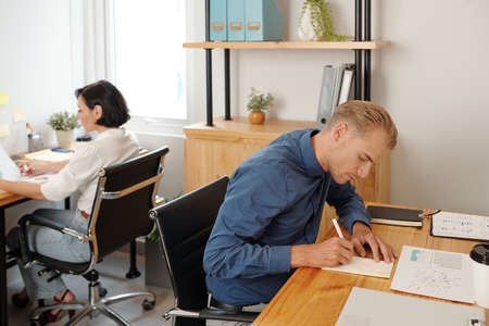 Serious concentrated young businessman writing his creative ideas and plans in planner when sitting at office deskの写真素材