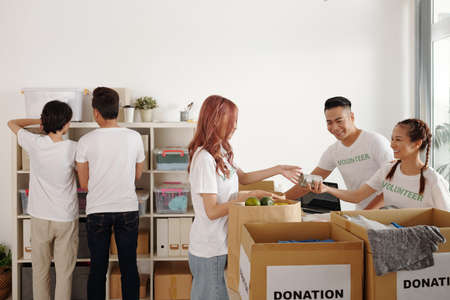 Group of young people working in community donation center, unpacking boxes and putting groceries and clothes on shelvesの写真素材