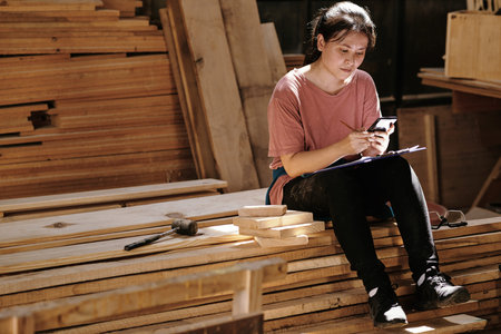 Young carpentry worker sitting on wooden planks reading text messages from customer and drawing...