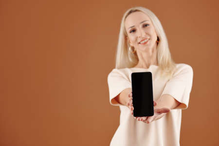 Positive young blond-haired woman in tshirt giving new smartphone with blank screen to you, studio shotの写真素材