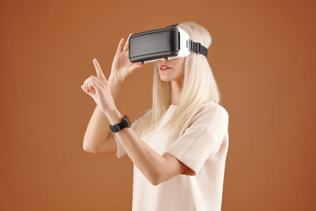 Modern curious young blond-haired woman in tshirt standing against brown background and touching...