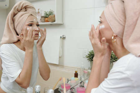 Serious young Caucasian woman with hair wrapped in towel smoothing cream on face skin in front of mirror in bathroomの写真素材