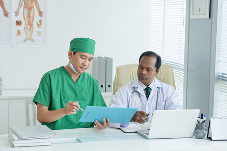 Serious general practitioner and his colleague discussing medical tests of patient when sitting at...