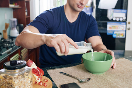 Close-up image of man sitting at kitchen counter and pouring milk in bowl of muesliの写真素材