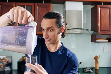 Emotional man pouring homemade protein cocktail in glass to drink before trainingの写真素材