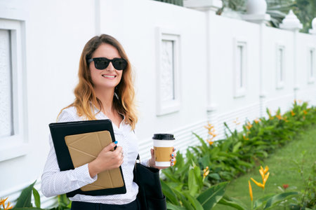 Happy elegant female entrepreneur standing outdoors with folder, tablet computer and cup of coffee