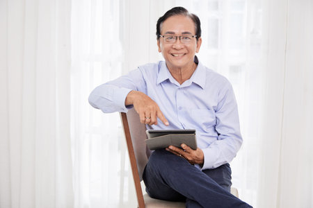 Positive elderly man sitting on chair and reading news or checking social media on tablet computer