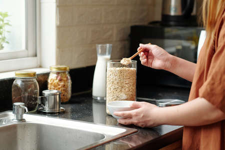 Woman standing at kitchen counter and putting spoon of quick oatmeal in bowlの写真素材