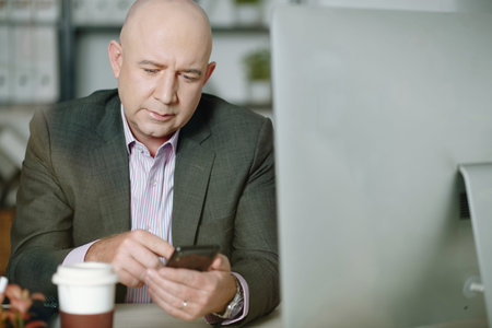 Senior businessman in suit sitting at his workplace in front of computer and typing a message on his...
