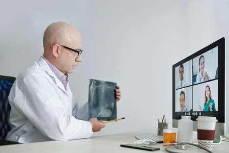 Mature doctor in white coat sitting at the table pointing at x-ray image and discussing it with his colleagues during online conferenceの写真素材