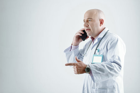Mature balded doctor in white coat talking on mobile phone isolated on white background