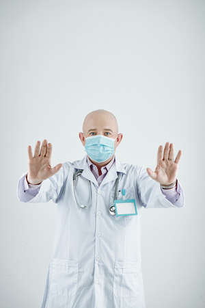 Portrait of bald mature doctor in mask showing his clean hands at camera standing against the white backgroundの写真素材