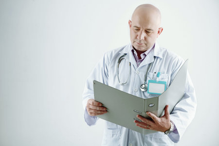 Mature doctor in white coat holding folder and examining medical card of his patient while standing...