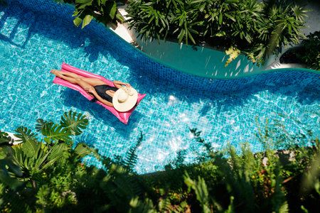 Beautiful young woman in black swimsuit and straw hat floating on pink mattress in swimming pool