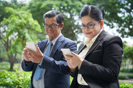 Positive senior business people in glasses standing in park and texting colleagues trying to manager work in office onlineの写真素材