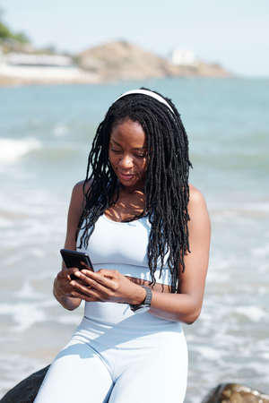 Fit young woman with smartphone resting on beach after training and checking text messages or setting relaxing musicの写真素材