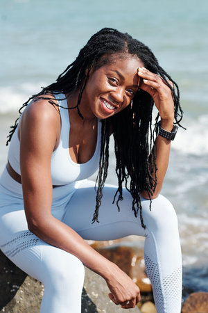 Portrait of cheerful pretty fit sportswoman resting on rock on beach after training and smiling at...