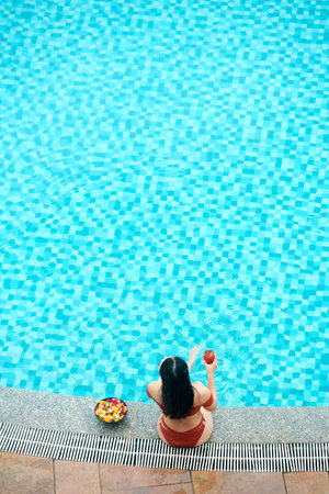 High angle view of black-haired woman in swimsuit drinking cocktail and eating fruits while relaxing...