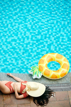 Unrecognizable woman in red swimwear sleeping with face covered summer hat near swimming pool