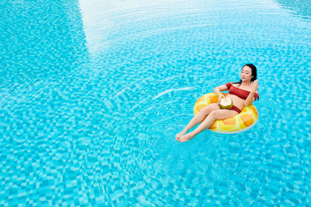 Young Asian woman in red swimwear sitting on inflatable ring and drinking coconut water in swimming...