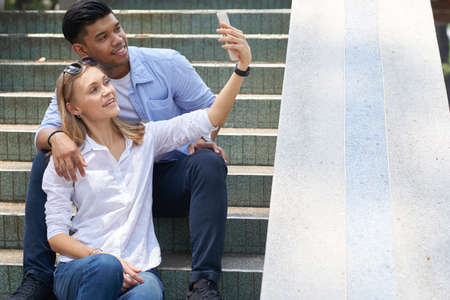 Happy diverse young couple sitting on steps and taking selfie to post on social mediaの写真素材