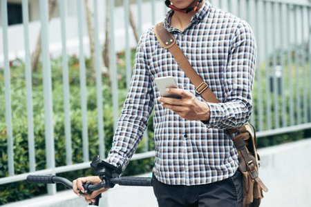 Cropped image of man in plaid shirt standing next to bicycle and checking notifications and...
