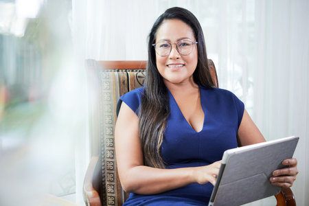 Portrait of cheerful mature elegant businesswoman in glasses sitting in chair and working on digital...