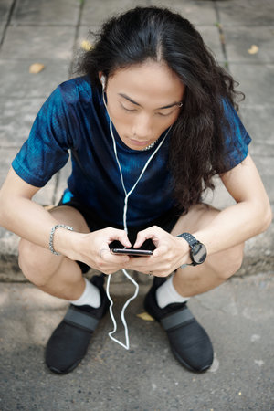 Serious young man sitting on pavement after morning run and answering text messages front friendsの写真素材