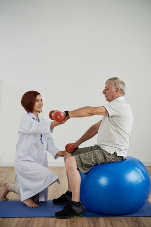Physical therapist asking senior man sitting on fitness ball to lift hand with dumbbell,...