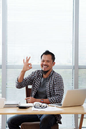 Portrait of happy exited entrepreneur in casual clothes sitting at office desk and showing ok signの写真素材
