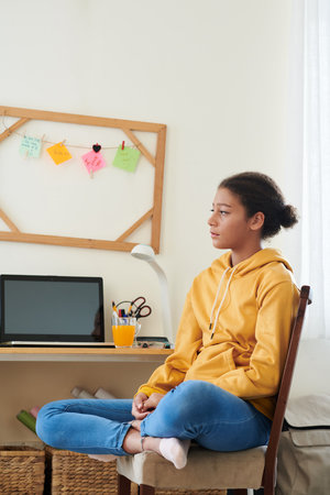Pensive bored schoolgirl sitting on chair at home waiting for online classes to startの写真素材