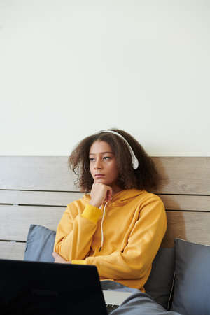 Pensive girl sitting in bed, listening to music in headphones and thinking how to solve coding problemの写真素材