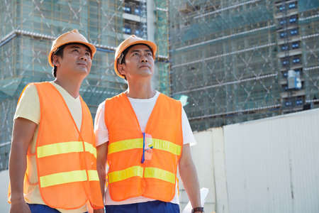 Team of builders in orange vests and had hats looking at high buildings of new hotel or business centerの写真素材