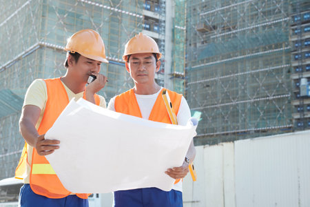 Contractor talking with builders via walkie-talkie when looking at building plan with head engineer...