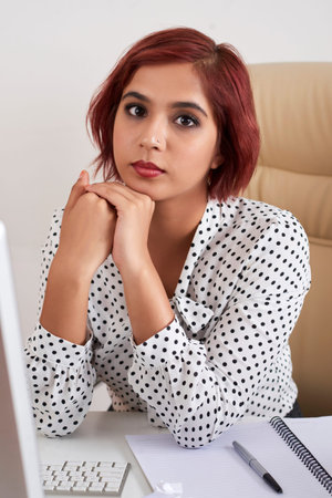 Serious young Indian businesswoman sitting at table with computer, documents and reportsの写真素材
