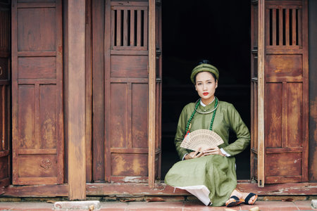 Beautiful young Vietnamese woman in traditional dress holding wooden fan when sitting on porch of...