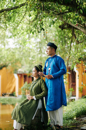 Beautiful just married Vietnamese couple in ao dai dresses sitting by pond and looking at waterの写真素材