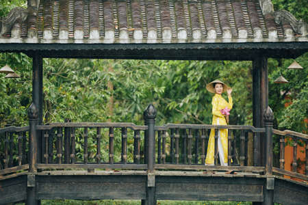 Smiling beautiful young Vietnamese woman in yellow dress and hat standing on bridge with bunch of lotus flowers and looking awayの写真素材