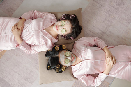 Joyful female friends relaxing with purifying clay mask and cucumber slices on face, view from aboveの写真素材