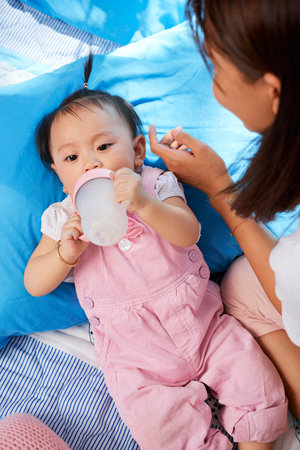 Young mother looking at lovely baby girl in pink overall drinking formulaの写真素材